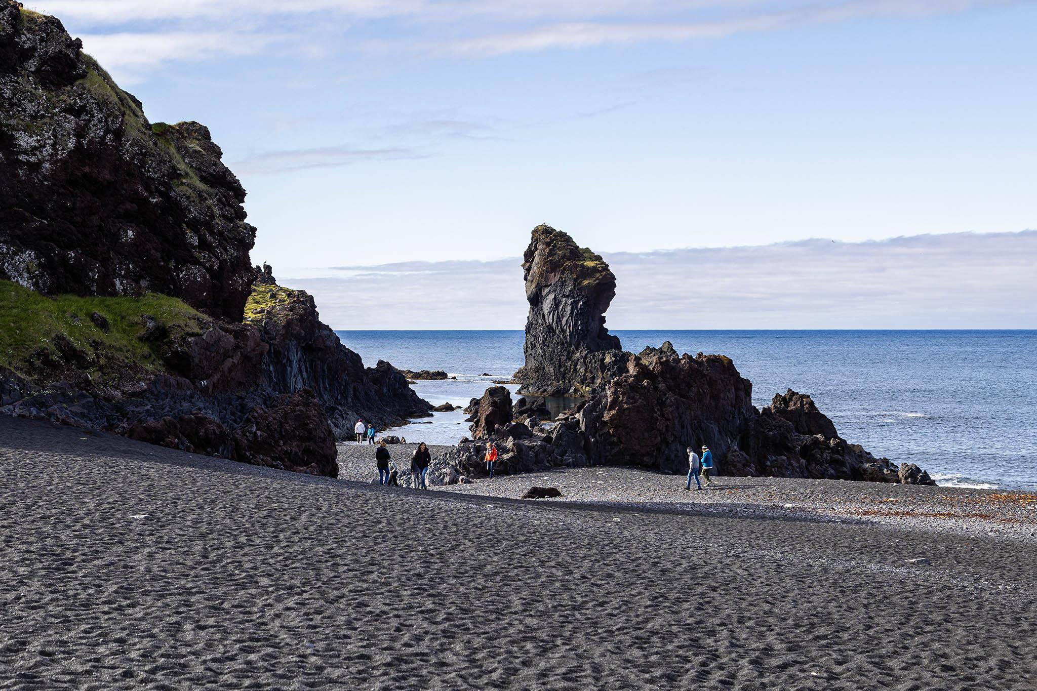 It's a cool day with a mostly clear sky except towards the far horizon over the sea where a light bank of cloud forms a wall and high up where some streamers of puffy white cloud stretch across the view. We're on a beach of black sand looking out to sea. The surface is dimpled from wind and footsteps and it looks soft and awkward to walk upon. From the left, by the flat water's edge there is a rocky rise of land. The rocky areas are largely black and volcanic-looking and where they're not sheer or where the crags angle back enough there is short green grass growing. The faces of the rock are dotted with white, possibly some form of moss or lichen but it's difficult to tell as we're quite some way back from the sea. Separate from the main mass of rock there's another rocky formation just at the tide line. It averages about five metres in height and twenty five metres in length on the beach but a section stretches out behind it into the shallow water where it then rises to a thick finger