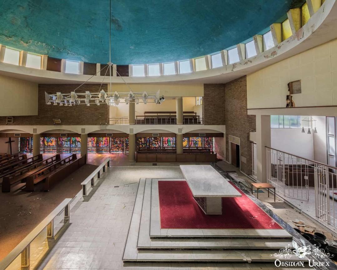 An overhead view of an abandoned church interior with a vast, peeling blue dome ceiling. Rows of wooden pews line the left, leading to an altar with a red carpet.