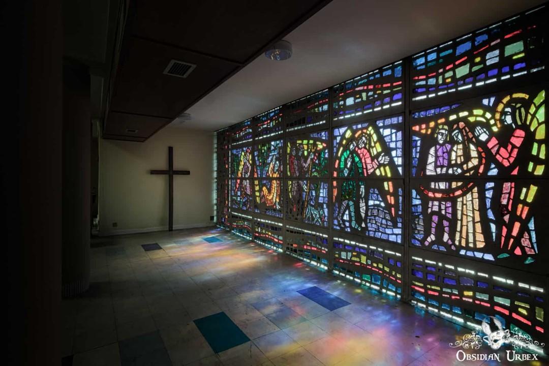 a vast, colorful stained-glass window on the right wall, casting vibrant patterns on the floor. A simple wooden cross hangs on the opposite wall.