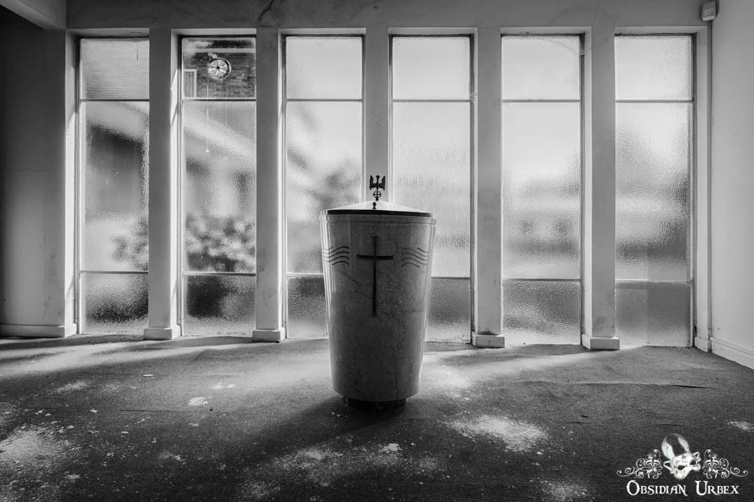A monochrome urbex photo of an abandoned church, featuring a baptismal font centered before tall, light-filled windows