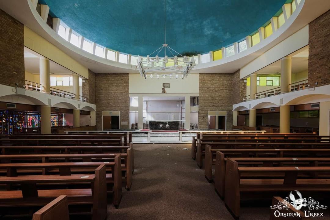 An abandoned church interior features rows of pews, a blue domed ceiling with a chandelier, and an altar area.
