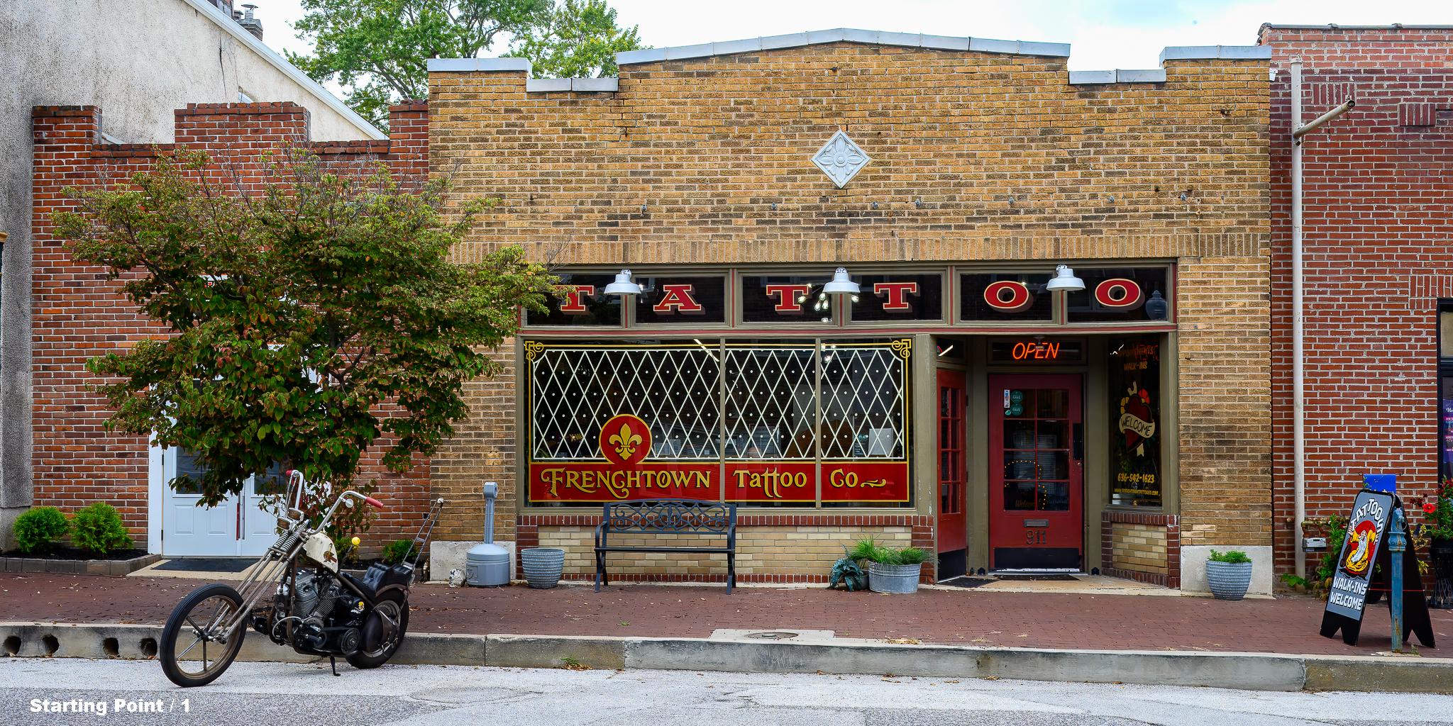 Front view of the Frenchtown Tattoo Company which is in a brick building.  There's an entrance door and 2 large windows plus 6 smaller windows over them.  A motorcycle is parked on the street in front of the building.