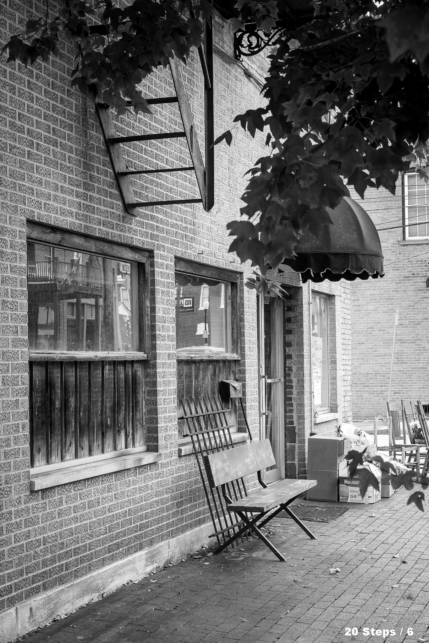 A brick building storefront.  There is a door with an awning over it and three windows.  The bottom of a fire escape is visible in the top left.  There is a bench and other items on the brick sidewalk.