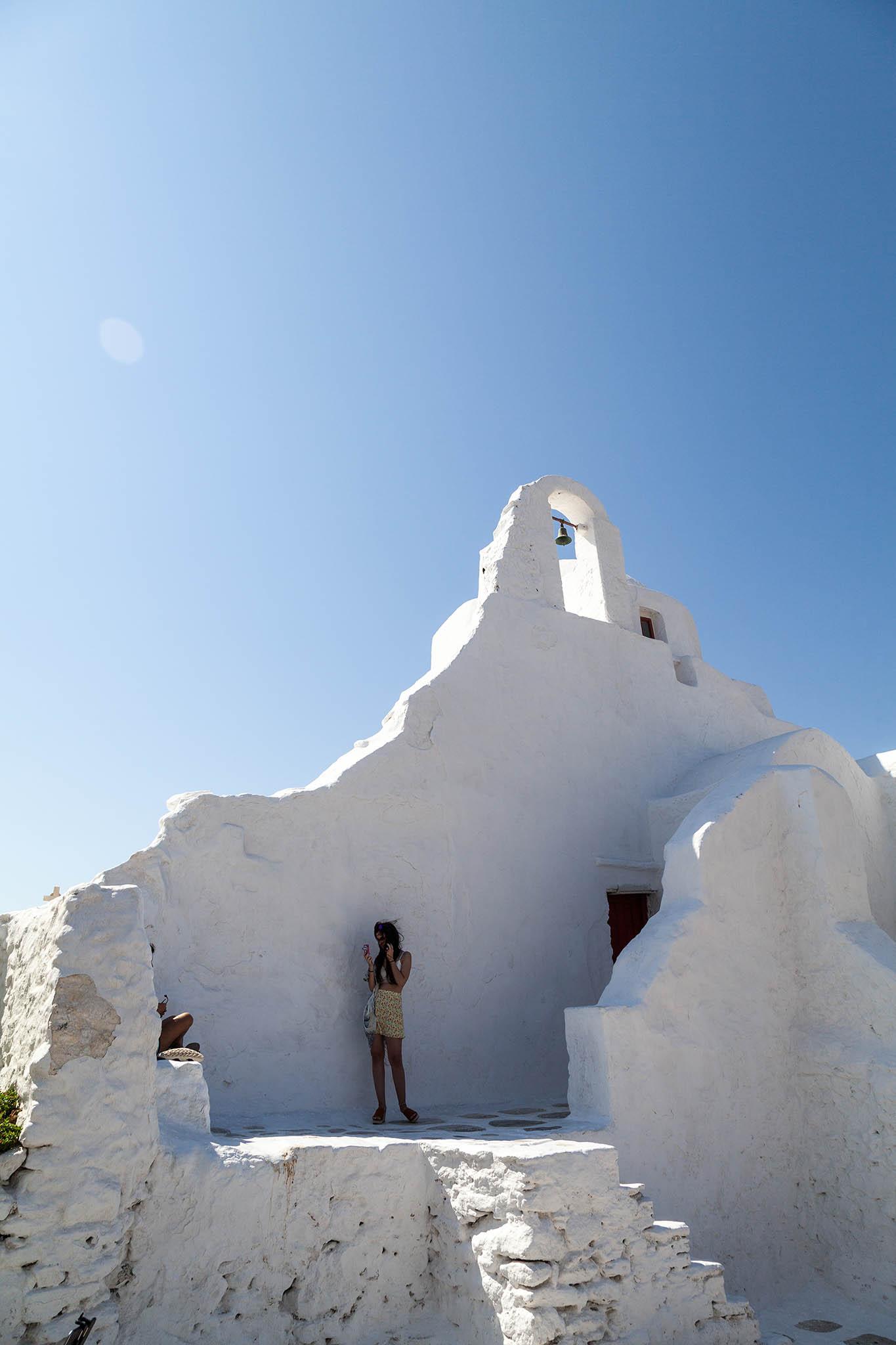 A minimalist shot in portrait orientation, broadly featuring just two colours. It's a hot, bright, sunny day with not a cloud in the piercing blue sky that brightens to a white-blue glow near the top left, close to where the sun is throwing a few lens flares into the scene. The bottom of the image contains a building, typical of Greek islands with its thick walls plastered and painted white. The building is a rough-contoured wall at the back of a small church with a set of steps up to a flat area that leads to a doorway barely visible behind another wall in front. Two young women are standing on the flat surface, one mostly behind a wall but clearly posing for her friend who's aiming a point-and-click camera at her. The wall of the church is about twice the woman's height where she's standing but it rises up twice that to a small niche that opens up and appears to hold a bell within its space.