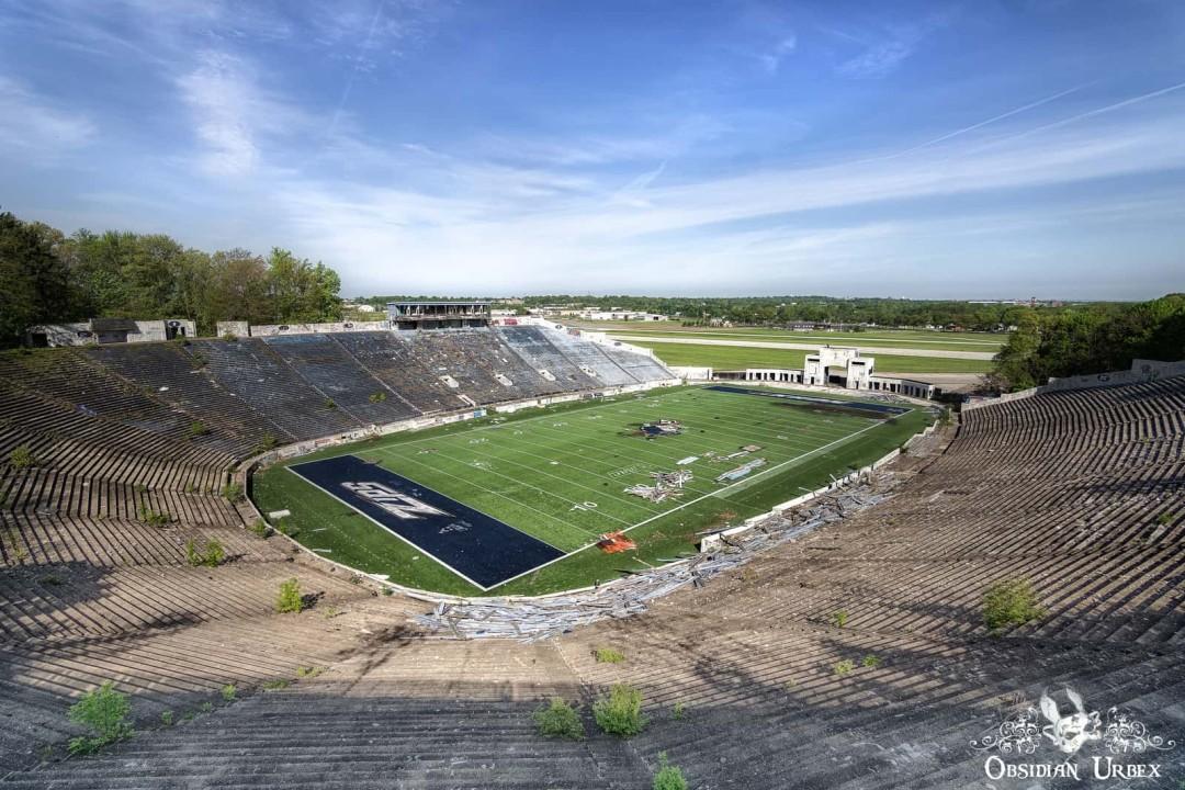 An aerial view of the abandoned Rubber Bowl stadium, featuring overgrown concrete stands and a football field still bearing the "ZIPS" logo