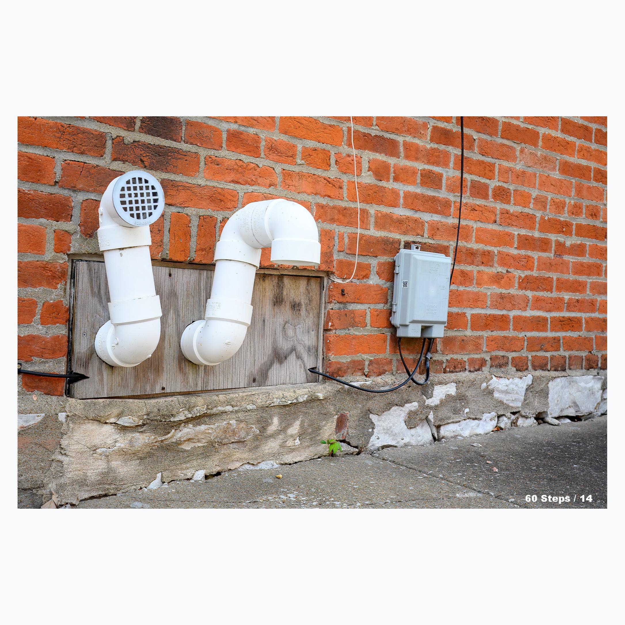 White PVC pipe vents coming out of the side of a brick building just above the sidewalk.