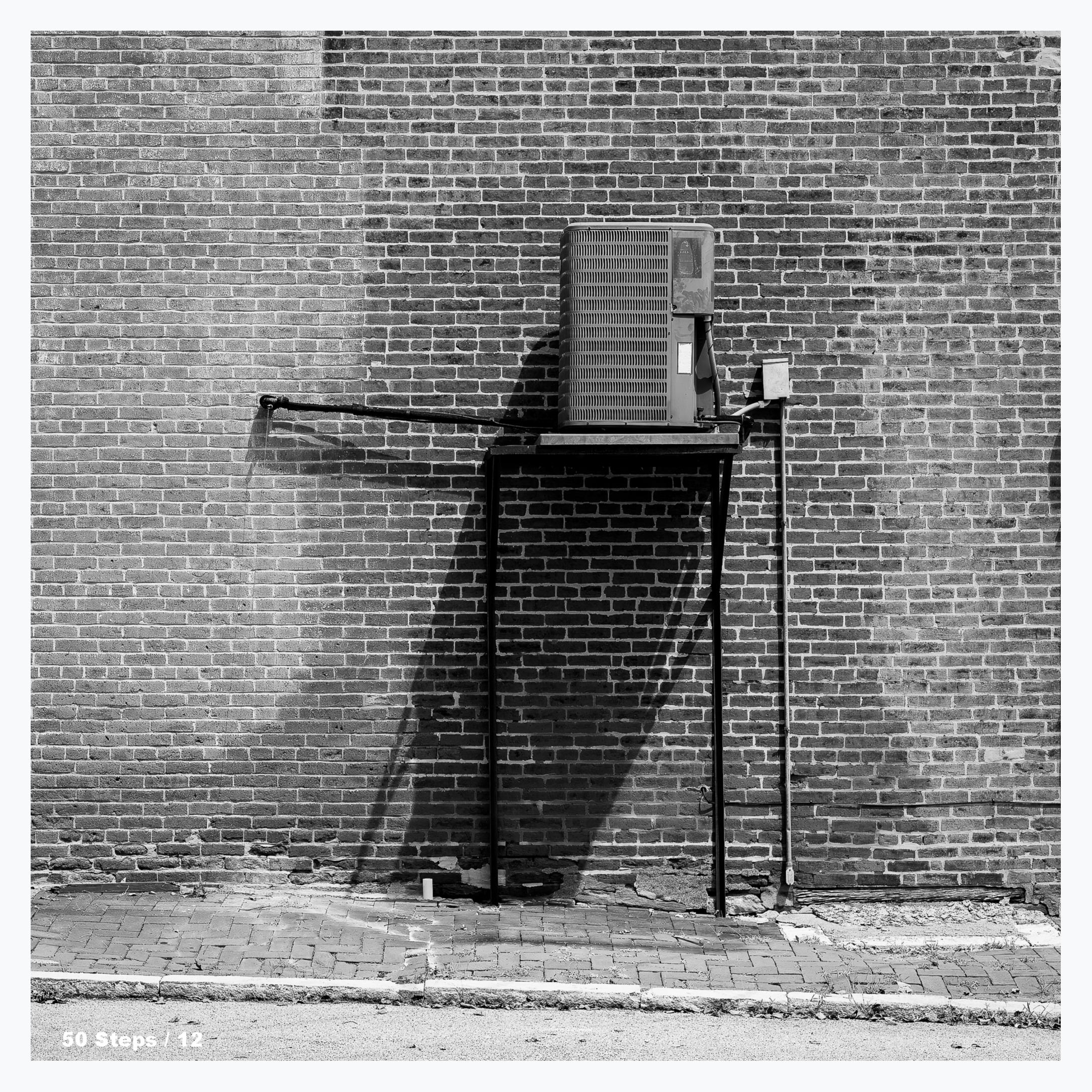 An A/C unit sits high above a sidewalk on metal supports.