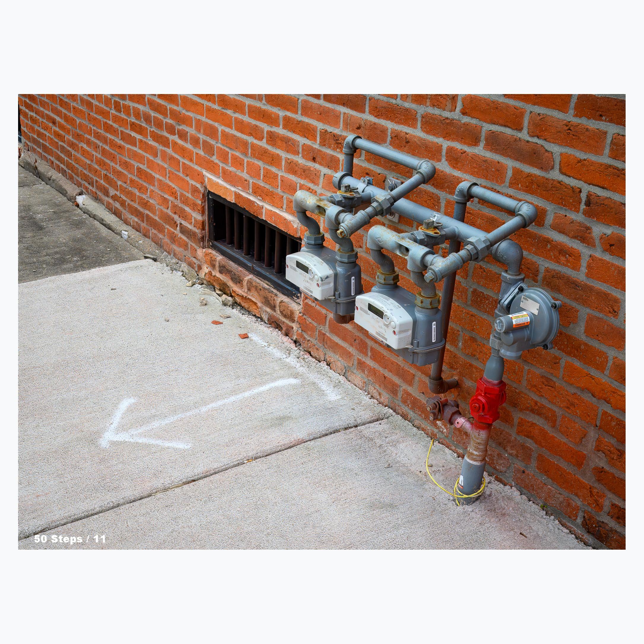 Gas meters and piping coming up from the sidewalk and then entering through the side of a brick building.