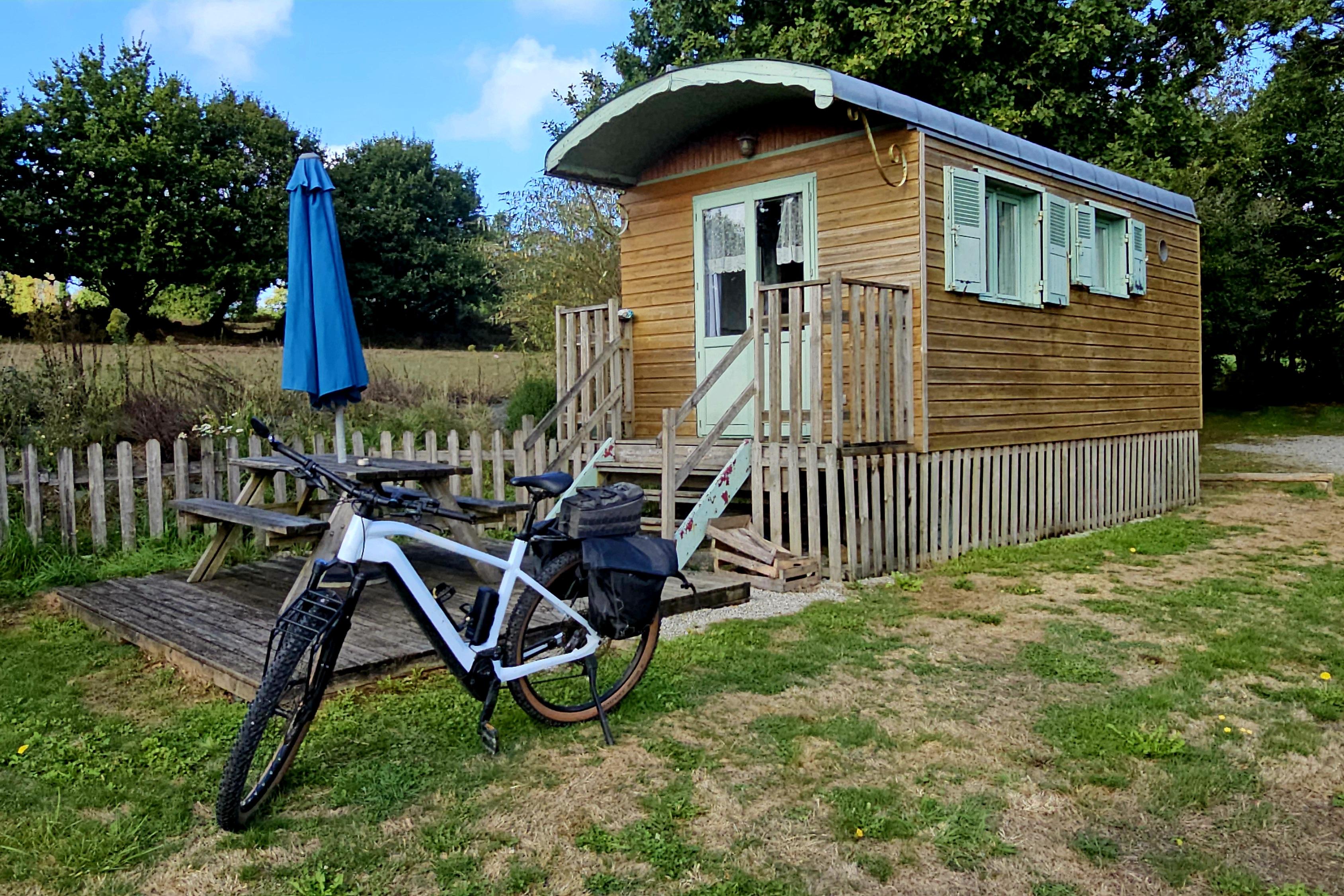 A wood-clad, caravan trailer sits in a field, with a bike in front of it.