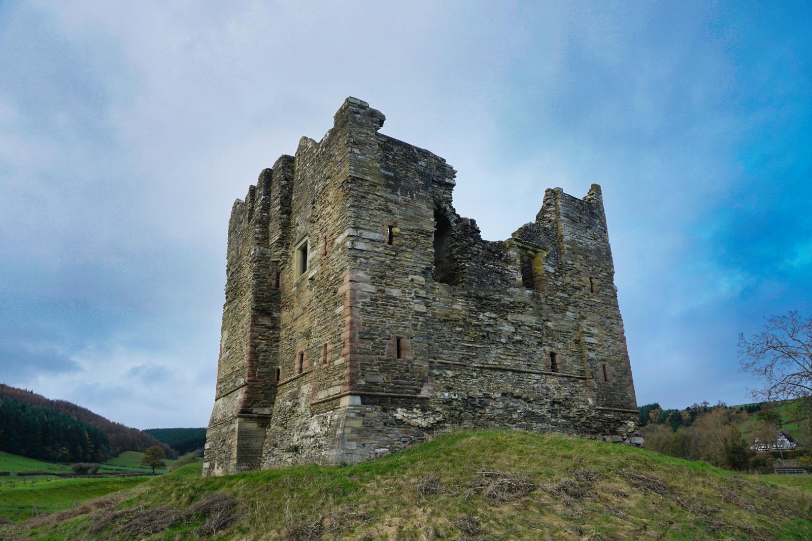 The ruins of Hopton Castle, Virtually cube shaped remains of the keep, with a large hole in one side. Several small slit like windows can be seen also
The remains stand on top of a small grassy mound with hills and trees in the background and a blueish cloudy sky above