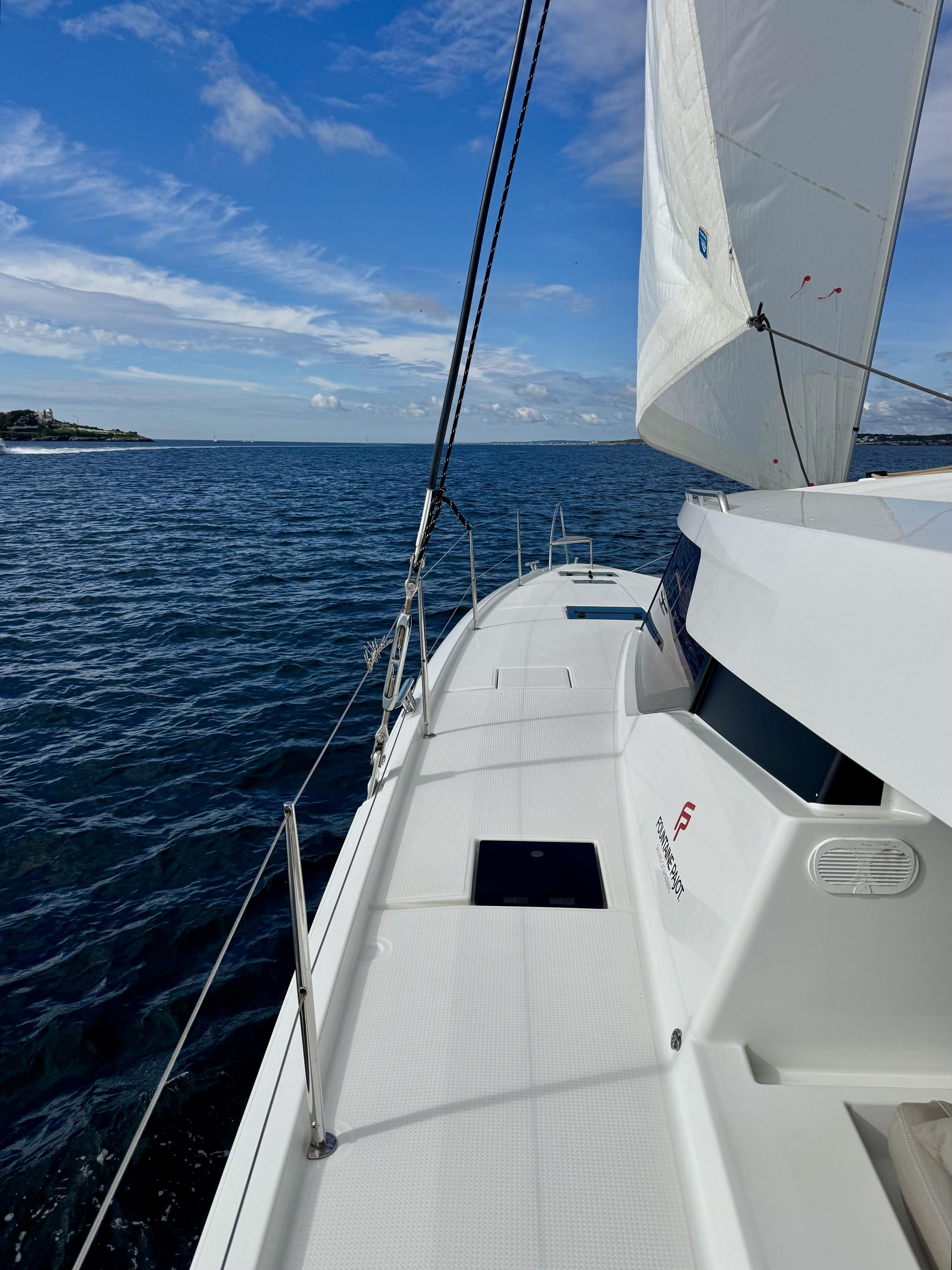 Photo taken on white sailboat looking forward toward horizon. Most of the left (“port”) side of boat is seen. The forward-most sail (“jib”) is also in view. A bit of land is seen on the left. Blue sky overhead with some puffy white clouds.

I took this photo while sailing out of Narragansett Bay, Rhode Island. The point of land on the left is Castle Hill, Rhode Island, which is one of the last bits of the bay (on this side) seen before entering the open ocean.