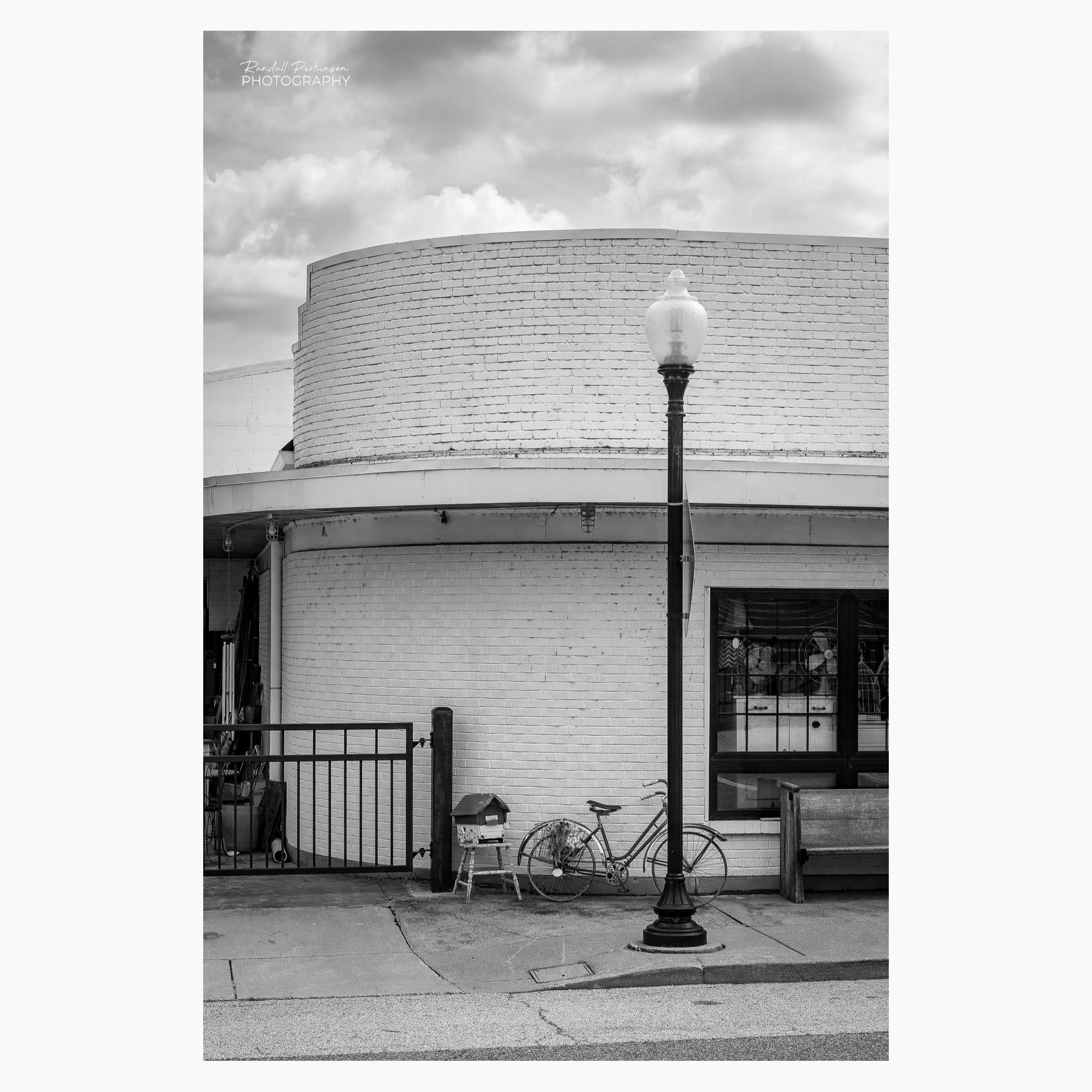 A one-story brick building, painted white, with a curved corner.  There is a bench, light pole, bicycle, and metal gate in front of the building.