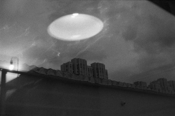 Ilford Delta 3200 (FF)

English
A black and white photograph of a high-rise residential building overlooked above a roof through a window. The reflection of a ceiling light and a lamp can be seen in the reflections.

中文
一张黑白照片.透过一个窗户的反光可以看到屋顶远处的一栋高层的住宅楼.窗户反光中映射出一个屋顶的灯和一盏台灯.