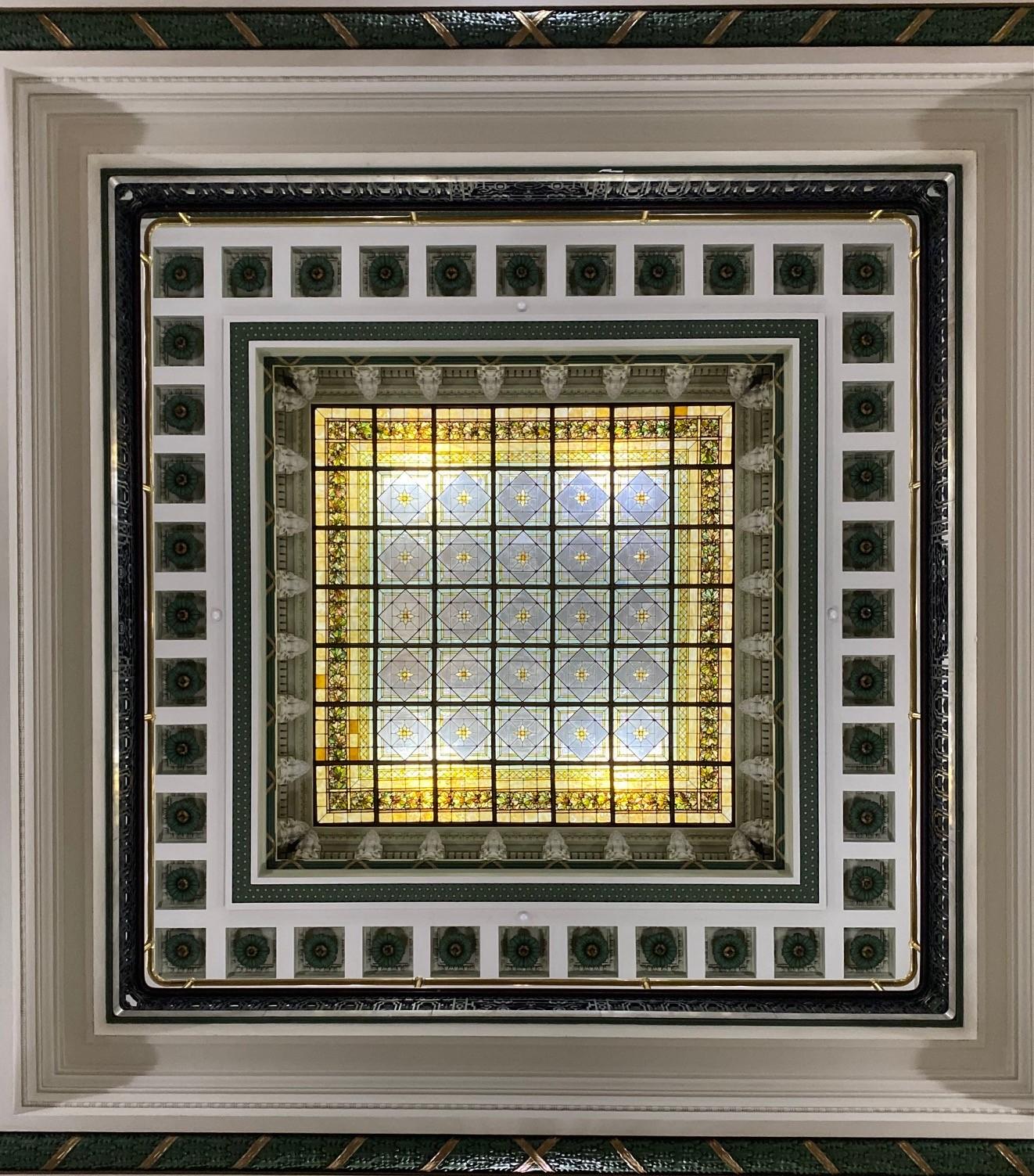 Color image, Monumental massive atrium skylight window, Summit County Courthouse circa 1908, Akron, Summit County, Ohio, USA. Image shows the complex geometric patterns in a massive stained-glass atrium skylight window at the top of the fourth story of the 1908 Courthouse building, flanked by geometric plaster casts corbels alternating in dark green and white, the panes and panels of the glass in gold, green, yellow, red, blue, and more, also arranged in a highly geometric pattern of squares within squares in a 7 x 7 repeating pattern, with 49 squares comprising the mains, with each individual of the 49 square panes having at least 16 separate panels, and, in many cases, far more in each of those panels.