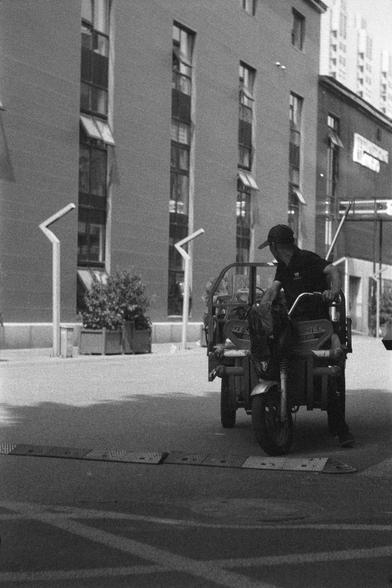 Ilford HP5 Plus 400 (FF)

English 
A black and white photograph. A person is driving a small tractor or truck in front of a multi-story building. The person is in the shadow cast by a tree.

中文
一张黑白照片.照片中一人在一栋高楼前驾驶着一辆三轮车.此人在树阴下.