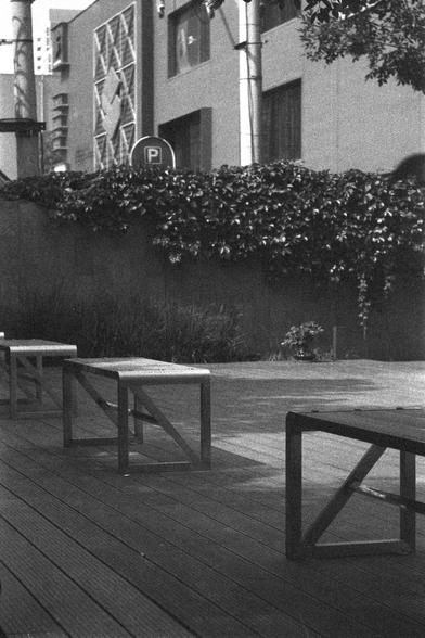 Ilford HP5 Plus 400 (FF)

English
A black and white photograph of a public space. Several benches stand in the shade. On the back of the space, a wall with crawling vines can be seen. Beyond the wall, a multi-story building and a parking sign can be seen.

中文
一张公共区域的黑白照片.几张长凳在阴影里.后面的墙上有植物藤蔓攀爬.背景中有一栋高层建筑和一个停车场的标志牌.