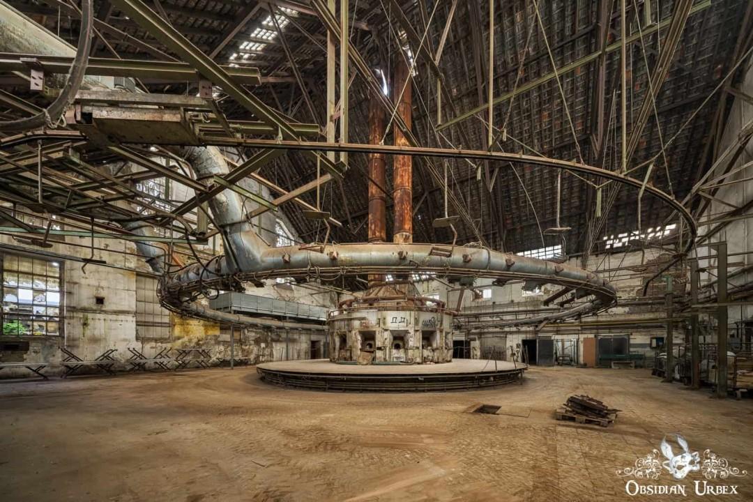 An abandoned industrial interior with a large circular machine marked "27 28." A suspended ring of pipes and beams encircles it under a high ceiling.