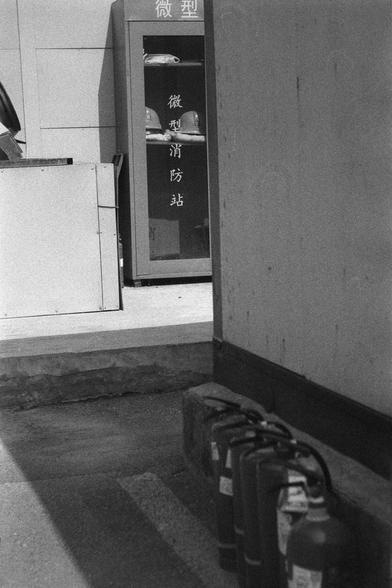 Ilford HP5 Plus 400 (FF)

English
A black and white photograph. Several fire extinguishers stand by the corner of the wall in the shade. A few meters away; there is a small cabinet with Chinese characters, reading Mini Fire Brigade, on them.

中文
一张黑白照片。照片中一些灭火器立在墙边的阴影中。几米开外有一个写着“微型消防站”的柜子。