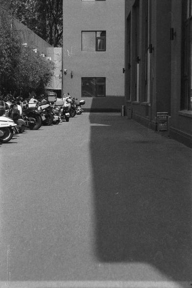 Ilford HP5 Plus 400 (FF)

English
A black and white photograph. The shadow of a high rise lies in the on the ground, separating the ground into two halves. The left is lit, with motorcycles parked in a line. The right side is a clear space.

中文
一张黑白照片。一栋高楼的影子在地面上制造出了一个分界线。左侧在阳光中，一排摩托车。右侧没有任何东西。