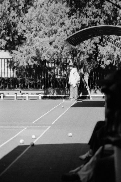 Lucky LUCKY SHD 400 (FF)

English
A black and white photograph of a group of people playing a ball game on the ground. The ground is painted with lines. A person is standing in the sunlight at the right corner of the lines. Some people are sitting on a bench in the foreground. Some trees are planted n the background.

中文
一张黑白照片. 一群人在玩一种球类游戏.地上画了白线.一群人坐在前景的阴影里.一个人站在阳光里线的角落.背景中有很多树.